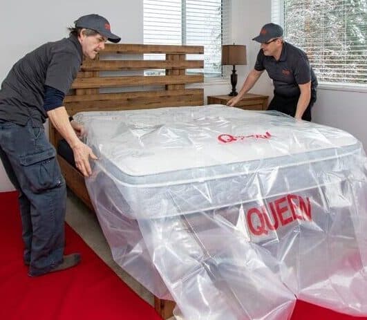 Two people lifting a wrapped queen-size mattress during a move.