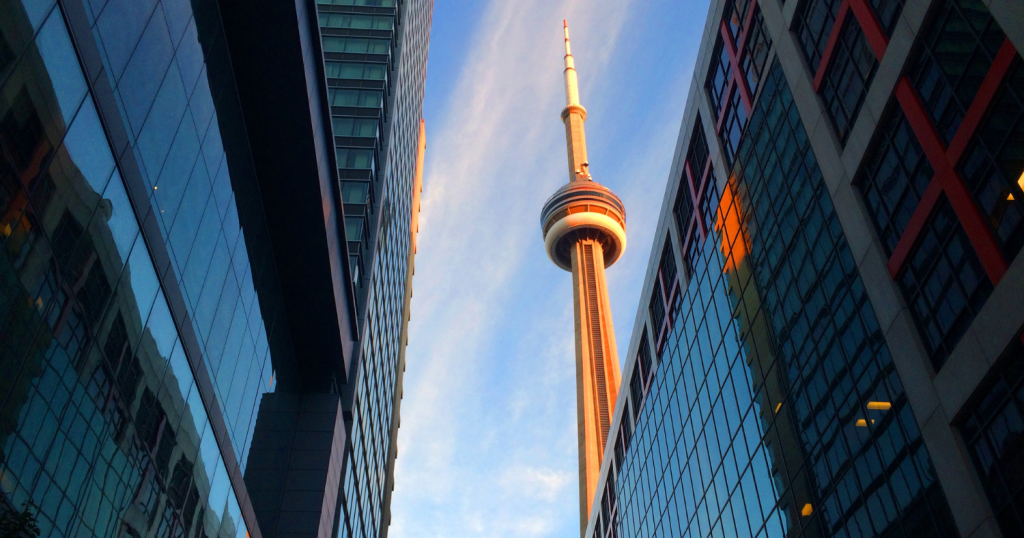The CN Tower framed by modern high-rise buildings in downtown Toronto, under a clear blue sky.