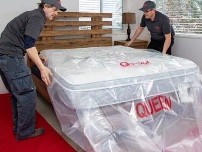 Two people lifting a wrapped queen-size mattress during a move.